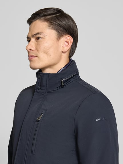 Männermodel trägt Funktionsjacke mit vielen Taschen von Geox in Dunkelblau, sichtbares Logo, glatte Struktur, Nahaufnahme Oberkörper.