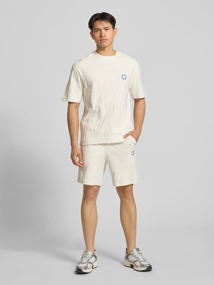 Männermodel trägt Regular Fit Shorts aus Frottee von HUGO in Creme mit Frottee-Optik, Tunnelzug, Logo-Stitching, seitliche Eingrifftaschen, aufgesetzte Tasche hinten, Ganzkörperansicht von vorne.