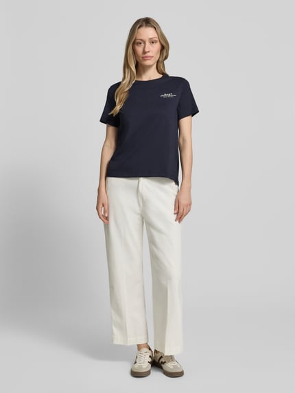 Frauenmodel trägt damski t-shirt z nadrukiem z logo i okrągłym dekoltem T-Shirt mit Label-Print und Rundhalsausschnitt von Gant, bawełna, krój regular fit, długość tyłu w rozmiarze S: 57 cm, kolor granatowy, proste białe spodnie, widoczne tekstury materiałów, Ganzkörperansicht von vorne.