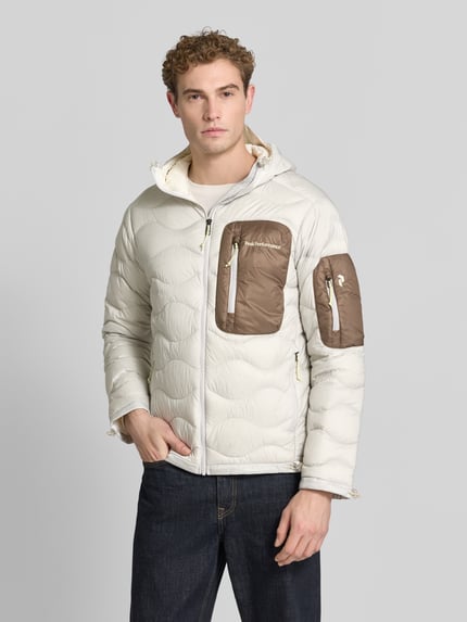 Męski model nosi Daunenjacke mit kontrastierender Brusttasche von Peak Performance, dwukolorowy, matowy, faktura, krój regular fit, długi, aplikacja: napis z logo, zamki błyskawiczne, kaptur, pasek do ściągania, kieszenie zapinane na zamek błyskawiczny, naszywana boczna kieszeń, kieszeń zapinana na zamek błyskawiczny, kształt kołnierza: kaptur. Ganzkörperansicht z przodu.