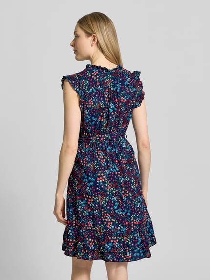 Modelka kobieca ma na sobie sukienkę mini z czystej wiskozy z kwiatowym nadrukiem na całej powierzchni Minikleid aus reiner Viskose mit floralem Allover-Print od Apricot, taliowany krój, dekolt w serek, bez rękawów, obszycie falbanami, listwa guzikowa, odpinana tasiemka w talii, długość tyłu w rozmiarze S: 89 cm, szerokość w rozmiarze S: 37 cm, kolorowa, lekka tkanina, widok całej sylwetki z tyłu.