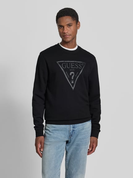Männermodel trägt Regular Fit Sweatshirt mit Logo-Detail Modell 'Audley' von Guess aus Baumwoll-Mix in Schwarz, Ganzkörperansicht von vorne. Rückenlänge 69 cm, Ärmellänge 69 cm.