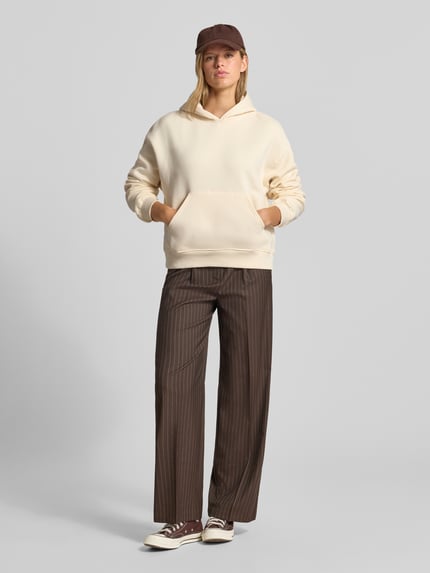 Modelka nosi Relaxed Fit Hoodie aus Baumwoll-Mix Modell 'Iconic' od Gina Tricot, beżowy kolor, kieszeń kangurka, elastyczne wykończenia, Ganzkörperansicht von vorne.