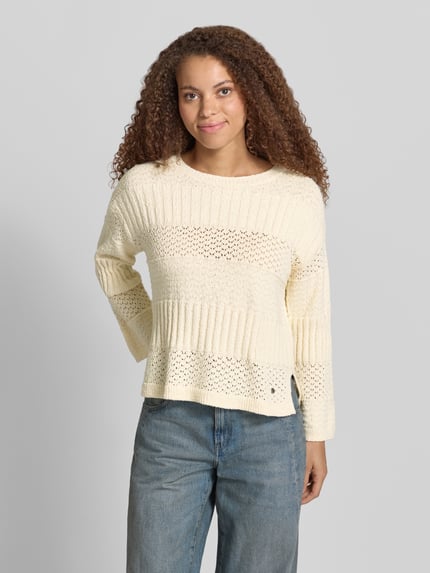 Frauenmodel trägt Boxy Fit Strickpullover aus reiner Baumwolle Modell 'ALAYA' von Pepe Jeans, Strickoptik, Struktur, Rundhals, Langarm, asymmetrisch, Materialverarbeitung Strick, Farbe Creme, ganzkörperansicht von vorne.