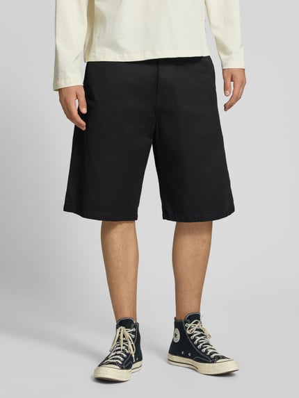 Männermodel trägt Bermudas mit Baumwoll-Anteil Modell 'Brader' von Carhartt Work In Progress, schwarzer Stoff, Relaxed Fit, Nahaufnahme Unterkörper, Produktdetails: Material-Mix, Knopf- und Reißverschluss, Eingriff- und Gesäßtaschen, Gürtelschlaufen, Bundweite 44 cm, Innenbeinlänge 30 cm. Ganzkörperansicht von vorne.