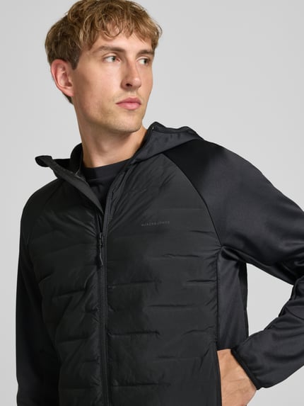 Männermodel trägt Hybrid Jacke mit Kapuze Modell 'PERFECT' von JACK & JONES in Schwarz, seitliche Reißverschlusstaschen, Nahaufnahme Oberkörper. Rückennaht und Logo-Print sichtbar, strukturierte Materialien, Kontext: Herrenjacke, gerader Schnitt, Reißverschluss.