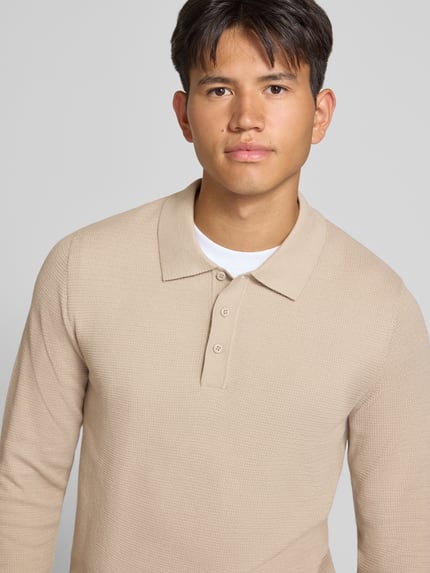 Męski model nosi koszulkę polo Slim Fit Poloshirt mit Label-Detail Modell 'AUSTIN' od JACK & JONES, mieszanka bawełny, krój slim fit, kołnierzyk polo, krój z długim rękawem, fakturowany wzór, detal z logo, długość tyłu w rozmiarze M: 69 cm, beżowy kolor, prosta faktura, zbliżenie na górną część ciała frontalnie.