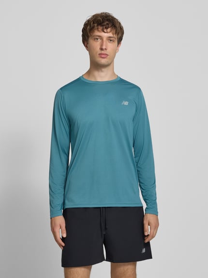 Męski model nosi Funktionsshirt mit Rundhalsausschnitt und Logo von New Balance, krój regular fit, bluzka z długim rękawem, okrągły dekolt, logo, Długość tyłu w rozmiarze M: 72 cm, widok całej sylwetki z przodu