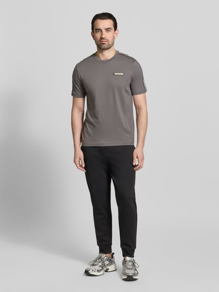 Männermodel trägt Regular Fit T-Shirt mit Logo-Patch von EA7 Emporio Armani in Grau, kombiniert mit schwarzer Hose, Ganzkörperansicht von vorne.