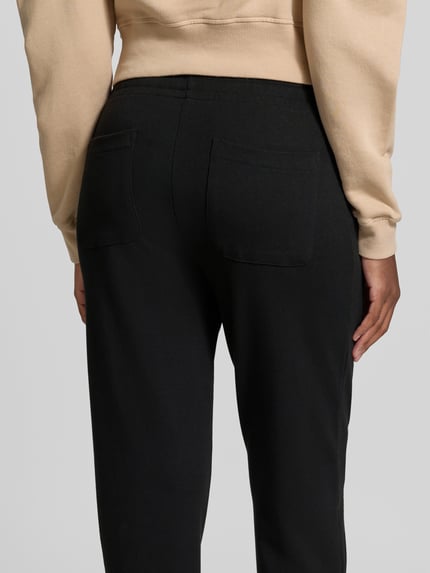 Modelka nosi spodnie dresowe Sweatpants in verkürzter Passform od Juvia, czarne, mieszanka bawełny i modalu, wpuszczane kieszenie, elastyczny pas, krój straight fit, skrócony, szerokość w rozmiarze S: 41 cm, długość wewnętrzna nogawki: 66 cm, widok całej sylwetki z tyłu.