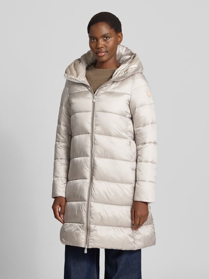 Frauenmodel trägt Steppmantel mit Kapuze Modell 'LYSA' von Save The Duck in Beige mit Zweiwege-Reißverschluss, Ganzkörperansicht von vorne.