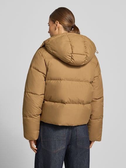 Frauenmodel trägt Pufferjacke mit Kapuze von LACOSTE in unifarbener Optik mit Logo-Stitching und Taschen, Ganzkörperansicht Rückansicht.