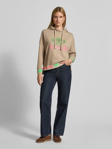 Frauenmodel trägt Oversized Hoodie mit Strasssteinbesatz Modell 'Namaste' von miss goodlife aus bawełna, krój oversized fit, kaptur z tunelem, nadruk z napisem, wykończenia: elastyczne, kolor beżowy z zielono-różowym nadrukiem, Ganzkörperansicht von vorne