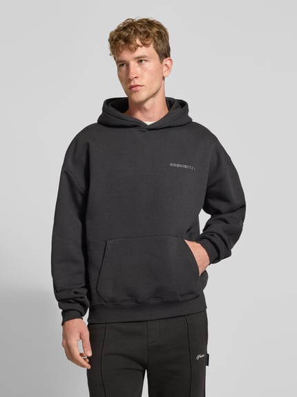 Männermodel trägt Relaxed Fit Hoodie aus Baumwoll-Mix mit Label-Detail von PROHIBITED, dunkler Farbton, Känguru-Tasche, elastische Abschlüsse, Ganzkörperansicht von vorne.