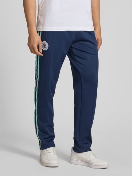 Mężczyzna nosi Regular Fit Sweatpants mit Vereins Logo Applikation Modell 'BLNACO' od adidas Originals, granatowy kolor, logo, kontrastowe paski, kieszenie, elastyczny pas z troczkiem, krój regular fit, widok na dolną część ciała z przodu.