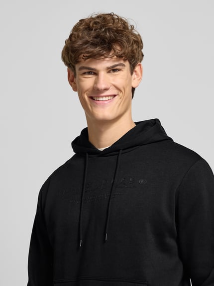 Männermodel trägt Hoodie mit Label-Print von Jack & Jones aus Baumwoll-Mix mit Kapuze, Tunnelzug, Känguru-Tasche, Lockeren-Schnitt, Nahaufnahme Oberkörper.