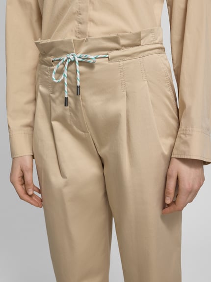 Frauenmodel trägt Straight Fit Stoffhose mit Tunnelzug von Marc Cain aus beigefarbenem Baumwoll-Mix mit gelegten Falten, Nahaufnahme Oberkörper.