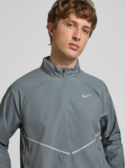 Männermodel trägt Regular Fit Jacke mit Stehkragen Modell 'Miler' von Nike Training in Grau mit Logo-Print, Reißverschluss, Nahaufnahme Oberkörper.