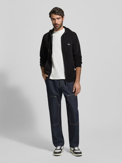 Männermodel trägt Sweatjacke mit Reißverschluss von LACOSTE, Baumwoll-Mix, Regular-Fit, Kapuze, Tunnelzug, Label-Detail, Eingrifftaschen, gerippte Abschlüsse, Rückenlänge 67 cm, Ärmellänge 65 cm, schwarze und blaue Farbtöne, glatte und strukturierte Oberflächen, Ganzkörperansicht von vorne.