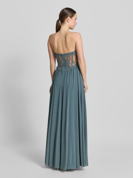 Frauenmodel trägt Abendkleid mit Spitze und Gehschlitz von Mascara in Blau, taillierter Schnitt, semitransparent, Rückansicht Ganzkörperansicht.