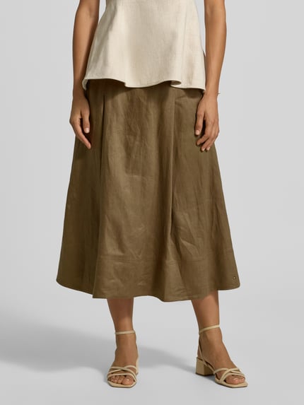 Kobieta nosi spódnicę midi z czystego lnu od Tommy Hilfiger, jednokolorowy efekt, rozkloszowana, zakładki w pasie, aplikacja z logo, zamek błyskawiczny, pasek, boczne wpuszczane kieszenie, materiał: len, widok na całą sylwetkę z przodu.