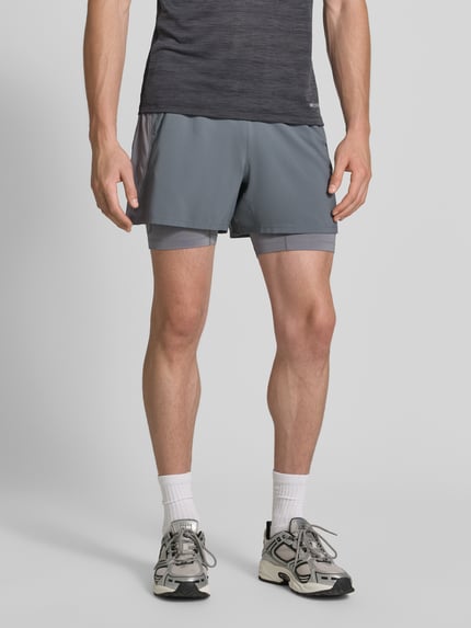 Männermodel trägt Regular Fit Shorts mit integrierten Tights Modell 'Road' von ASICS TIGER in Grau, elastischer Bund, Logo-Detail, Ganzkörperansicht von vorne.