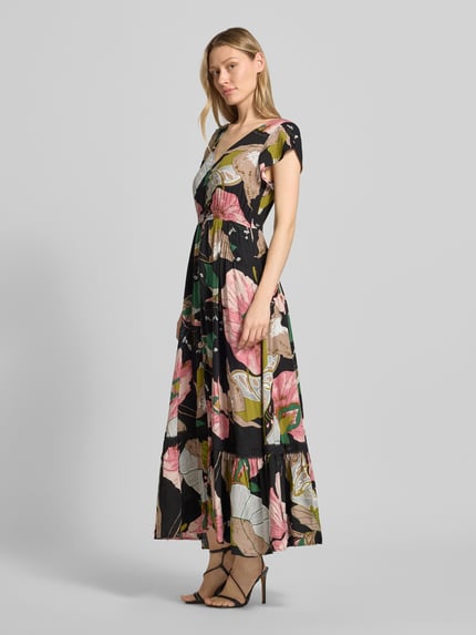 Vrouwenmodel draagt Maxikleid aus Viskose mit V-Ausschnitt van Apricot, zwart met kleurrijk bloemenpatroon, zuiver viscose, getailleerd, V-hals, volants, steekzakken, ruglengte maat S 130 cm, borstwijdte 43 cm, mouwlengte 11 cm, zijaanzicht, bijna volledige lichaamsopname.