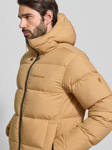 Männermodel trägt Steppjacke mit Kapuze und Label-Stitching von PEAK PERFORMANCE in Beige, sichtbare Eingrifftaschen, Reißverschluss, Nahaufnahme Oberkörper. Regular-Fit, Herren, Rückenlänge 72 cm, Schulterbreite 49 cm, Brustweite 52 cm, Ärmellänge 70 cm.