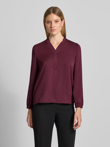 Kobiece model nosi Regular Fit Bluse mit V-Ausschnitt od Marc Cain, krój regular fit, dekolt w serek, detal z logo, długość tyłu w rozmiarze 36: 65 cm, długość rękawów w rozmiarze 36: 59 cm, kolor bordowy, gładka faktura, widok całej sylwetki z przodu.