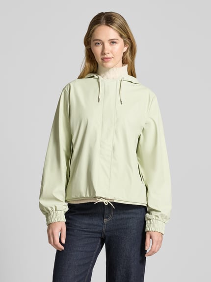 Vrouwenmodel draagt Jacke mit Abdeckleiste van RAINS in lichtgroen, rechte fit, tunnelkoord aan kap en zoom, elastische mouwuiteinden, detailopname bovenlichaam.
