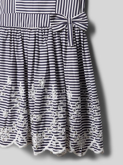 Mädchen trägt Regular Fit Kleid mit floralem Stitching von DANAMADE aus Reine Baumwolle, Lochmuster, Zierschleife, Nahaufnahme Unterteil.