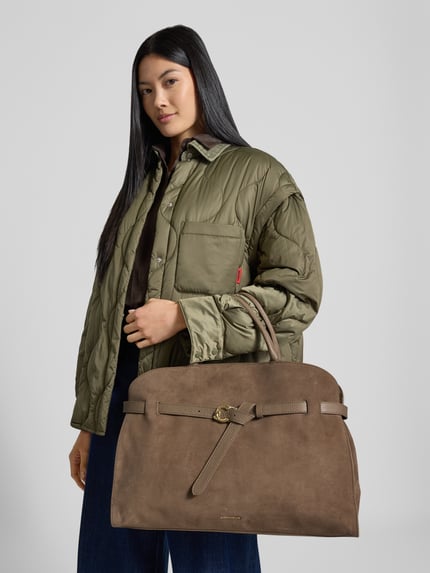Frauenmodel trägt Handtasche mit Logo-Detail Modell 'Sabine' von Coccinelle in Braun, sichtbar genarbte Textur, Ganzkörperansicht von vorne. Geteiltes Hauptfach, Magnetverschluss, verstellbarer Schulterriemen, Innenfach mit Reißverschluss, Maße: 44x32x16 cm, Schulterriemen 106 cm.