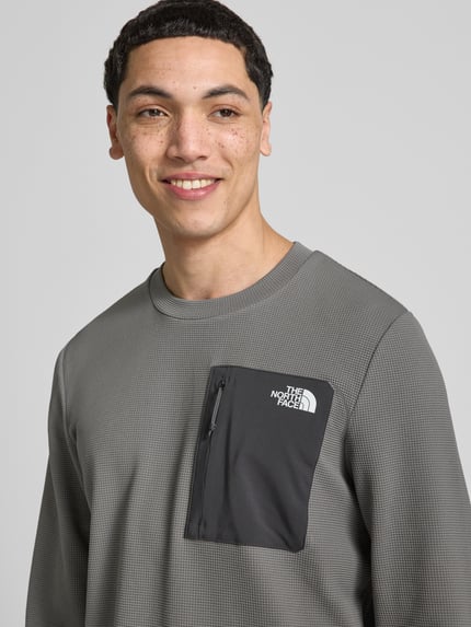 Männermodel trägt Langarmshirt mit Napoleontasche Modell 'KECHA' von The North Face, Elastischer-Material-Mix, Label-Detail, Nahaufnahme Oberkörper.