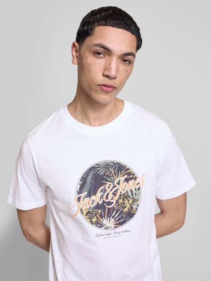 Męski model nosi t-shirt z nadrukiem Jack & Jones T-Shirt mit Rundhalsausschnitt Modell 'HONOLULU' z bawełny, krój regular fit, okrągły dekolt, długość tyłu 68 cm w rozmiarze M, jasne kolory, nadruk z przodu, gładka tekstura tkaniny. Ganzkörperansicht z przodu.