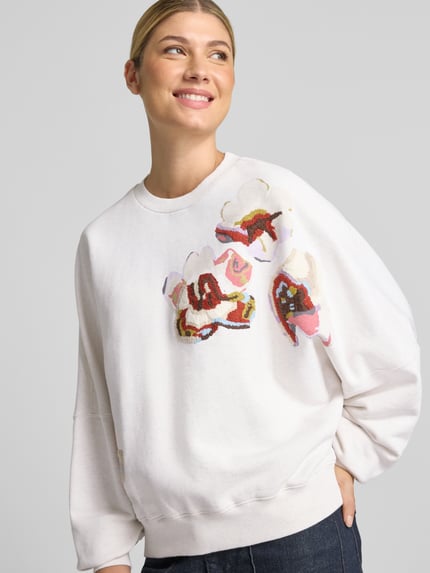 Vrouwenmodel draagt oversized sweatshirt uit puur katoen Model 'ASTERA' van DRYKORN, ribboorden, bloemmotief, witte en roze tinten, nahaufnahme bovenlichaam.