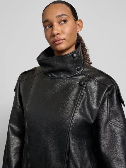 Frauenmodel trägt Kunstlederjacke mit hohem Stehkragen Modell 'ONLVANI' von ONLY in schwarzer Lederoptik mit Druckknöpfen und Schulterklappen, sichtbare Nähte und Gummizug-Details, unifarben, Nahaufnahme Oberkörper.