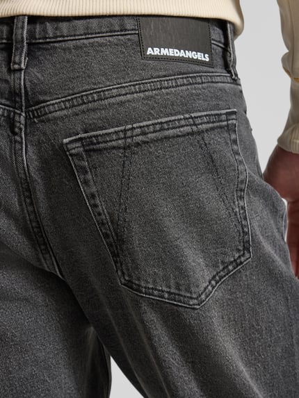 Männermodel trägt Tapered-Fit Jeans aus Bio-Baumwoll-Mix Modell 'MAAKX' von Armedangels in grauer Denim-Optik mit Label-Patch und 5-Pocket-Design, Detailaufnahme Rückansicht. Kontext: Herren Tapered-Fit Jeans Passform Relaxed Tapered-Fit, Bundhöhe Mid, Gürtelschlaufen, Knopf und verdeckter Reißverschluss, Material Bio-Baumwoll-Mix.