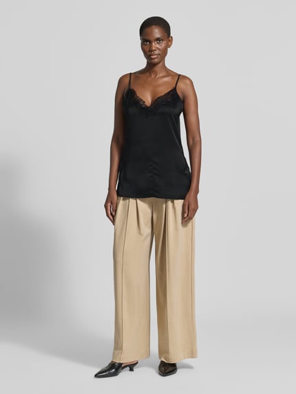 Frauenmodel trägt schwarzes Spitzentop mit V-Ausschnitt Modell 'Brenda' von My Essential Wardrobe aus Satin mit Spitzenbesatz, Regular Fit, verstellbare Spaghettiträger, kombiniert mit beiger weiter Hose, Ganzkörperansicht von vorne.