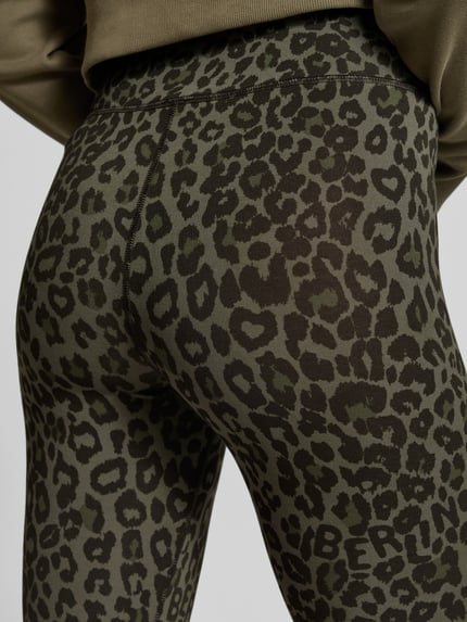 Frauenmodel trägt Leggings aus Viskose-Mix von Lala Berlin mit Leopardenmuster in Oliv und Schwarz, Nahaufnahme Rückansicht. Eng anliegend, elastischer Bund, High Waist, knöchellang.