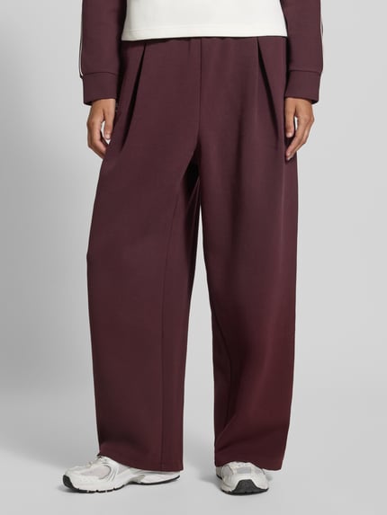 Frauenmodel trägt Regular Fit Sweathose aus Baumwoll-Mix von Lacoste Sport in Dunkelrot mit weitem Bein und glatter Textur, Nahaufnahme Unterkörper.