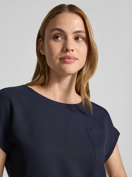 Frauenmodel trägt Blusenshirt mit verlängertem Rücken und Brusttasche von monari, dunkelblauer Stoff, Rundhalsausschnitt, Aufschlag an kurzen Ärmeln, Brusttasche, Label-Detail, Gerader-Schnitt, Damen, Nahaufnahme Oberkörper.