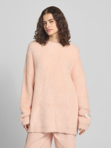 Frauenmodel trägt Oversized Strickpullover mit Label-Detail von Karo Kauer in hellem Rosa, grobe Textur, Rundhalsausschnitt, überschnittene Schultern, Ganzkörperansicht von vorne.