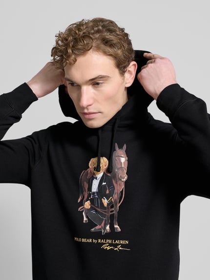 Männermodel draagt Hoodie met Labelprint van Polo Ralph Lauren in zwart katoenmix, los model, capuchon met tunnelkoord, ribboorden, nahaufnahme oberkörper.