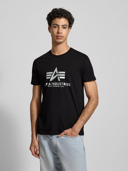 Männermodel trägt T-Shirt mit Label-Print von Alpha Industries, Schwarz, Reine Baumwolle, Gerader Schnitt, Rundhalsausschnitt, Ganzkörperansicht von vorne.