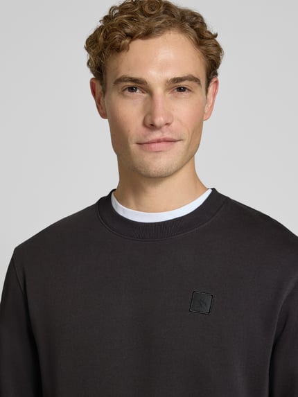 Męskamodel nosi Regular Fit Sweatshirt aus reiner Baumwolle mit Label-Patch von Scotch & Soda, kolor czarny, naszywka z logo, krój regular fit, bawełna, wykończenia: elastyczne, długość tyłu w rozmiarze M: 70 cm, długość rękawów w rozmiarze M: 66 cm, zbliżenie na górną część torsu.