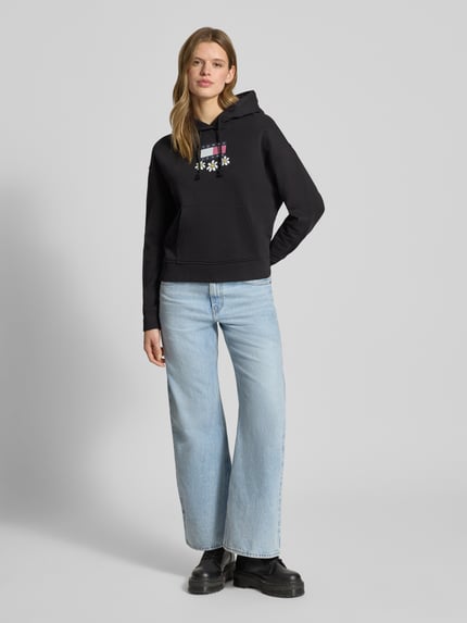 Vrouwenmodel draagt Hoodie mit Label- und Motiv-Print Modell 'DAISY FLAG' van Tommy Jeans, zwart, losse fit, unidesign, katoenmix, geribde boorden, kangoeroezak, vooraanzicht hele lichaam.