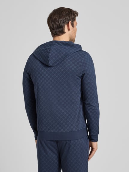 Mannelijk model draagt 'Sweatjacke mit Logo-Muster und Kapuze' van JOOP! Collection in donkerblauw katoen, regular fit, met logopatroon, capuchon, rits, steekzakken, ruglengte maat M 69 cm, mouwlengte 63 cm. Ganzkörperansicht rückansicht.