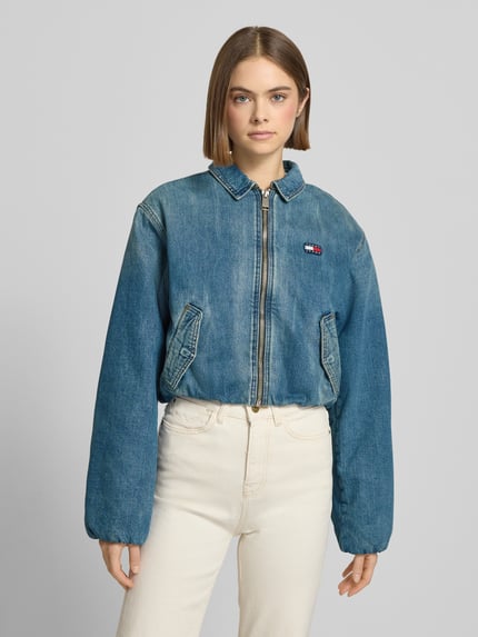 Kobiece model prezentuje Regular Fit Jeansjacke aus reiner Baumwolle von Tommy Jeans, niebieski denim, zapięcie na zamek, boczne kieszenie, aplikacja z logo, widok całej sylwetki z przodu.