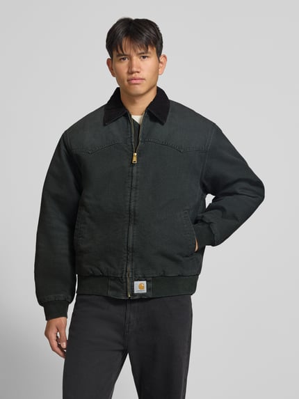 Männermodel trägt Jacke mit Reißverschluss und Label-Detail Modell 'Santa Fe' von Carhartt Work In Progress in dunklem Baumwollstoff, Ganzkörperansicht von vorne. Regular Fit, Umlegekragen, sichtbarer Zipper.