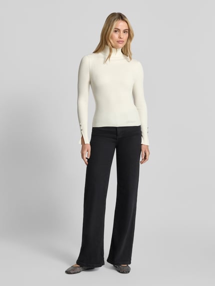 Kobiece modelka nosi Strickpullover aus Viskose-Mix mit Rollkragen od GUESS, krój slim fit, model 'ELLIE', rippowana faktura, detal z logo, kolor kremowy, długość tyłu 54 cm, długość rękawa 62 cm, widok całej sylwetki z przodu.